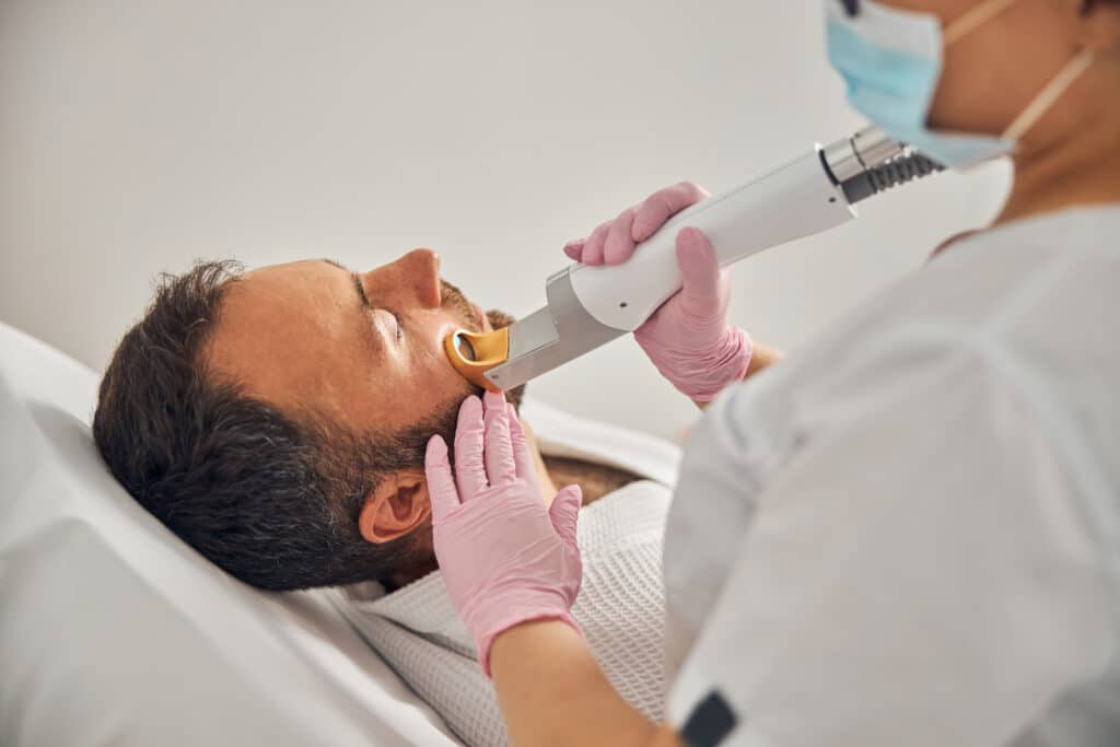cerca de un joven apuesto acostado en el divan mientras la cosmetologa elimina el vello no deseado de la mejilla con un dispositivo laser - Dani